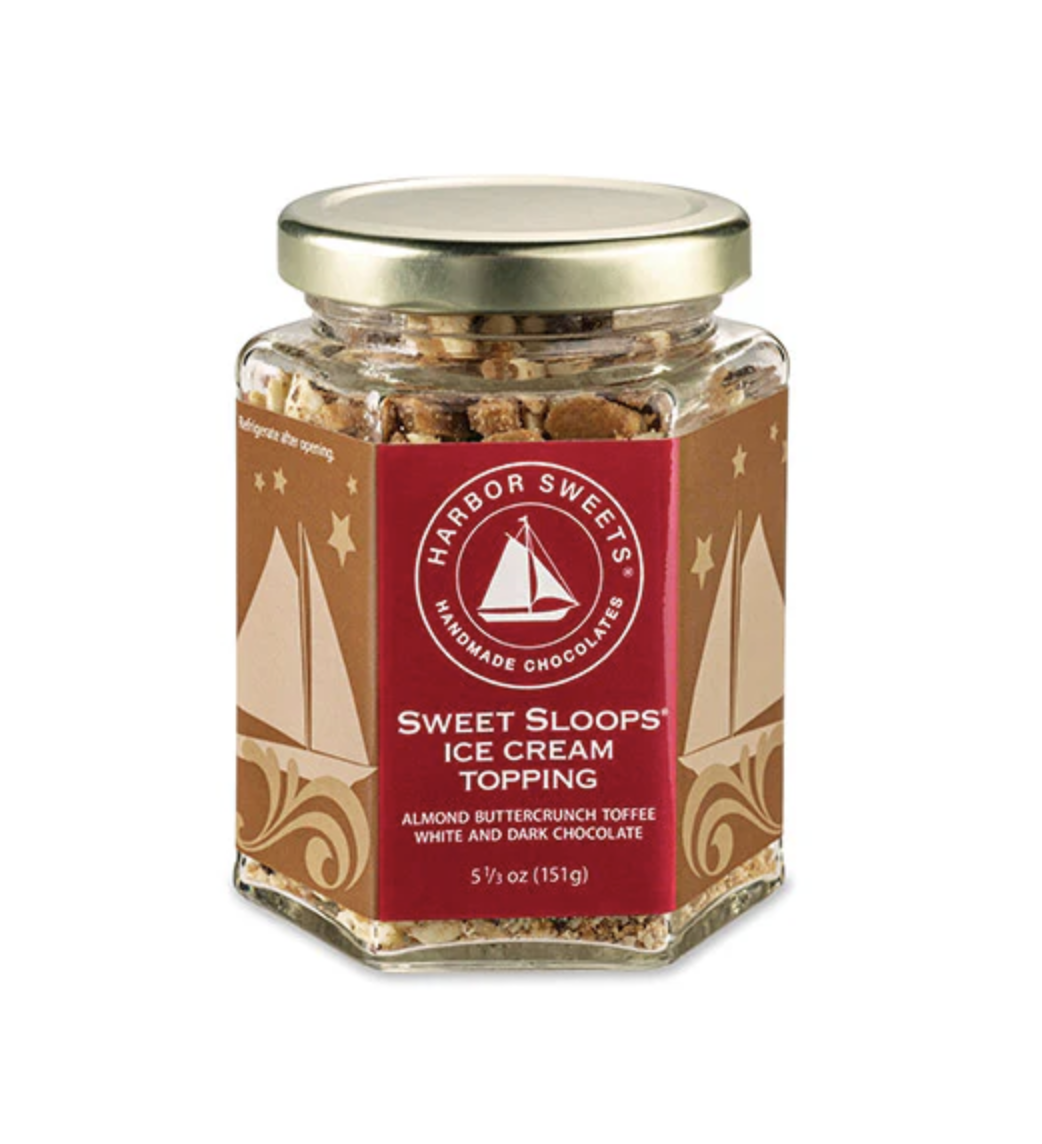 A hexagonal glass jar of Sweet Sloops Ice Cream Topping — almond buttercrunch toffee with white and dark chocolate. The label wraps the jar in warm brown with a red Harbor Sweets sailboat seal, gold metal lid, and decorative sailboats illustrated along the sides.
